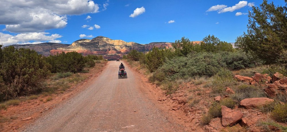 A rugged off-road vehicle navigates the winding trails of Sedona's iconic Red Rock Country, kicking up a cloud of dust as it climbs over rocky terrain. Dramatic sandstone formations in shades of red, orange, and yellow rise up in the distance, their jagged silhouettes framed against a brilliant azure sky. Warm, directional sunlight casts deep shadows, accentuating the curves and contours of the landscape. The vehicle's driver leans forward, focused and determined, as they tackle the challenging path ahead, surrounded by the captivating natural beauty of this remarkable wilderness oasis.
